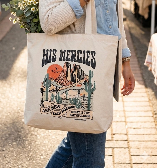 A person wearing a cream-colored canvas tote bag featuring a printed desert landscape illustration with cacti, mountains, a sun, and the text "His mercies are new each morning, great is thy faithfulness.