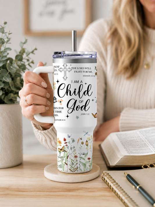 A white stainless steel tumbler with a handle featuring "I Am A Child of God" text, a cross, and botanical illustrations, held by a person sitting at a desk with an open Bible
