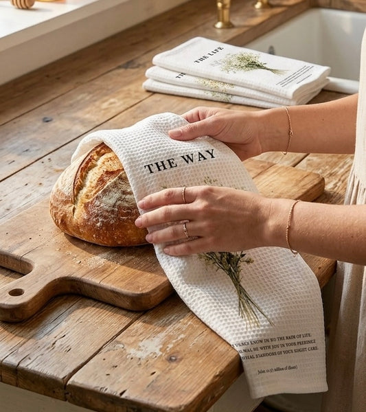 pair of hands holding a white waffle-weave kitchen towel printed with a bouquet of flowers and the text "THE WAY" over a loaf of artisan bread on a wooden kitchen counter
