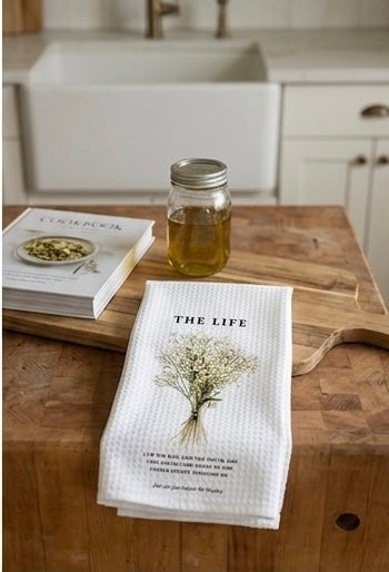 A white waffle-weave kitchen towel featuring a bouquet of dried baby's breath and the text "THE LIFE" laid on a rustic wooden cutting board next to a glass jar and a cookbook.