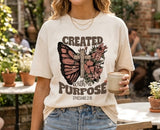 A woman smiling, wearing an aesthetic cream-colored Christian t-shirt featuring a floral butterfly cross and the scripture Ephesians 2:10