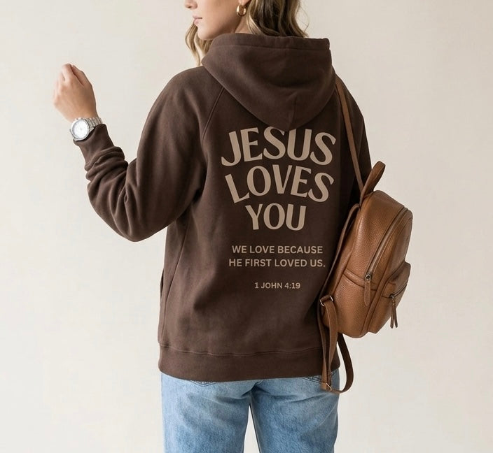 Woman wearing a brown oversized hoodie with 'Jesus Loves You' and '1 John 4:19' printed in large, modern typography on the back