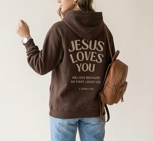 Woman wearing a brown oversized hoodie with 'Jesus Loves You' and '1 John 4:19' printed in large, modern typography on the back
