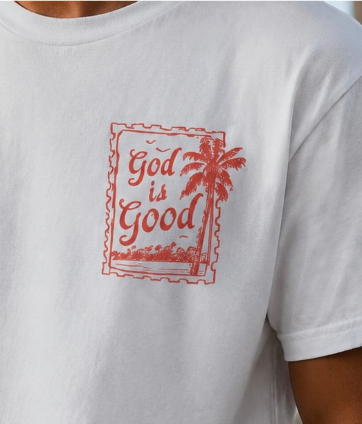 Detailed close-up of a red "God is Good" palm tree stamp design printed on a white faith-based Christian graphic t-shirt.