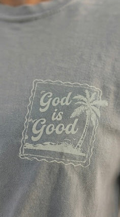 Close-up detail of a sage green Christian t-shirt featuring a minimalist "God is Good" palm tree stamp design on the chest.