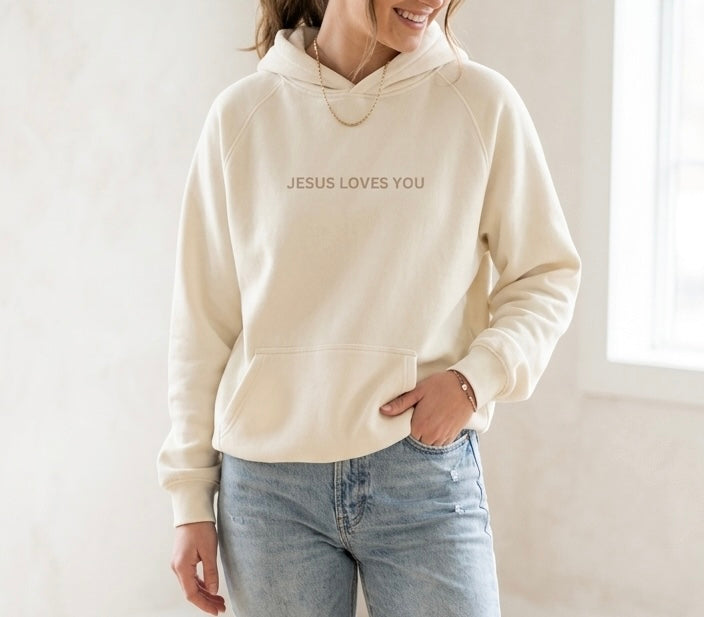 Minimalist cream hoodie featuring 'Jesus Loves You' in elegant sand-colored text on the chest, worn by a woman