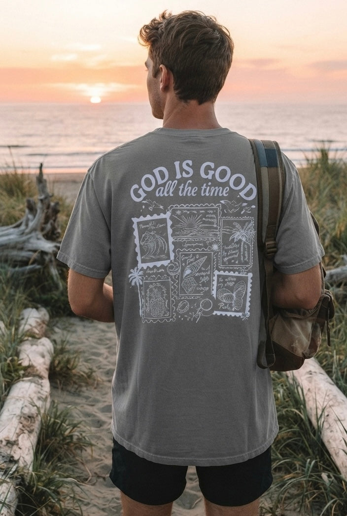 Man wearing a sage green inspirational Christian t-shirt with a detailed "God is Good all the time" back print, standing on a beach at sunset.