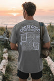 Man wearing a sage green inspirational Christian t-shirt with a detailed "God is Good all the time" back print, standing on a beach at sunset.