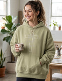 A woman wearing a Christian baggy, light green hoodie with a cross design on the front, holding a coffee mug in an art studio