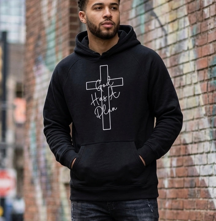 A man wearing a black "God Has A Plan" hoodie with a cross design, styled in an urban setting against a brick wall