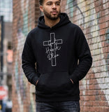 A man wearing a black "God Has A Plan" hoodie with a cross design, styled in an urban setting against a brick wall
