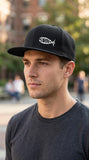 Side profile of a young man wearing a black Christian snapback hat with a white Jesus fish logo in a casual urban setting. Modern men's faith clothing and lifestyle appare