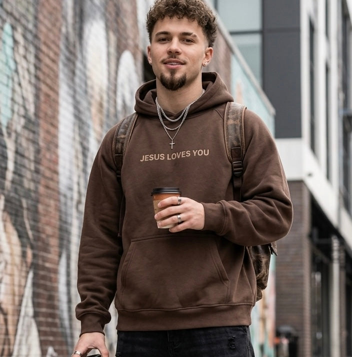 Young man wearing a brown 'Jesus Loves You' minimalist Christian streetwear hoodie, styled with dark cargo jeans and sneakers in an urban setting.