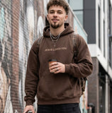Young man wearing a brown 'Jesus Loves You' minimalist Christian streetwear hoodie, styled with dark cargo jeans and sneakers in an urban setting.