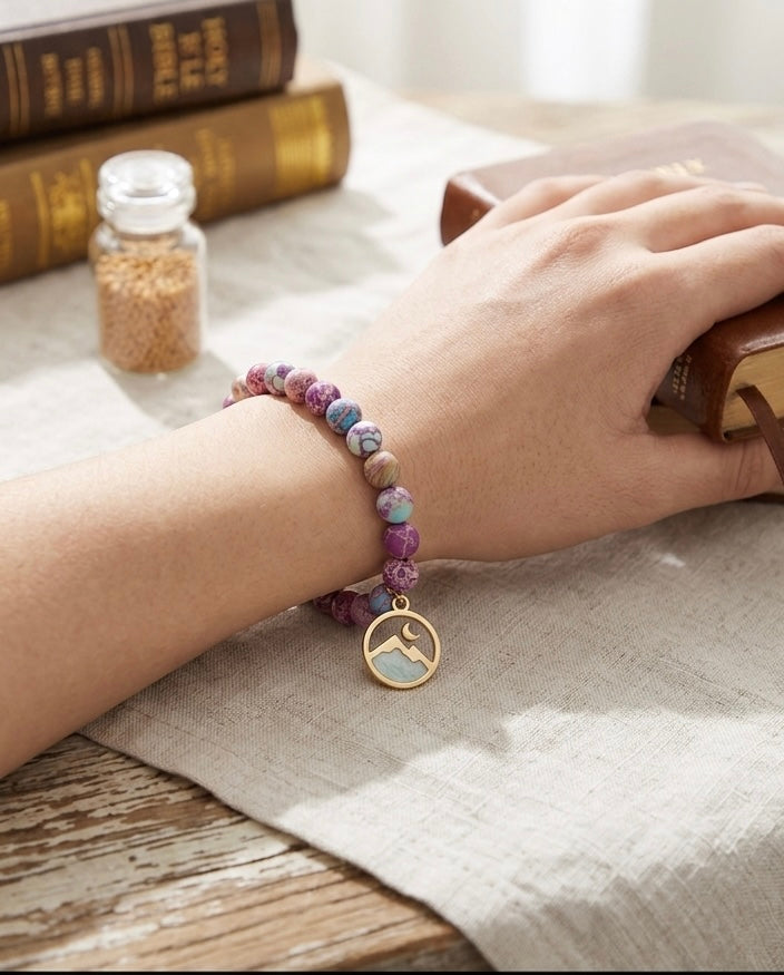 A woman's hand wearing a purple marble beaded bracelet with a mountain charm, resting on a table next to a Bible and a small jar of mustard seeds, perfect as a Christian faith-based gi