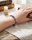 A woman's hand wearing a purple marble beaded bracelet with a mountain charm, resting on a table next to a Bible and a small jar of mustard seeds, perfect as a Christian faith-based gi