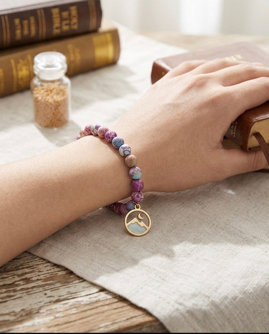 A woman's hand wearing a purple marble beaded bracelet with a mountain charm, resting on a table next to a Bible and a small jar of mustard seeds, perfect as a Christian faith-based gi