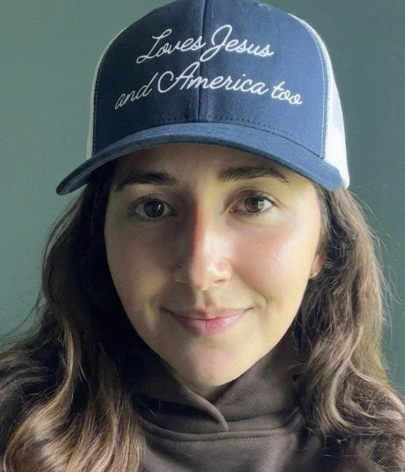 Woman wearing a navy blue Christian faith-based trucker hat with Loves Jesus and America too embroidery.
