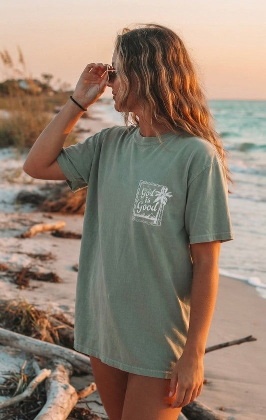 Woman wearing a sage green Christian apparel t-shirt with a faith-based "God is Good" palm tree stamp design on the beach