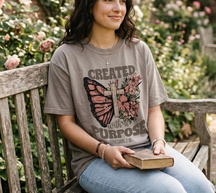 A woman sitting on a garden bench with a Bible, wearing a brown Christian t-shirt with a floral butterfly cross design and Ephesians 2:10 text