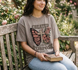 A woman sitting on a garden bench with a Bible, wearing a brown Christian t-shirt with a floral butterfly cross design and Ephesians 2:10 text