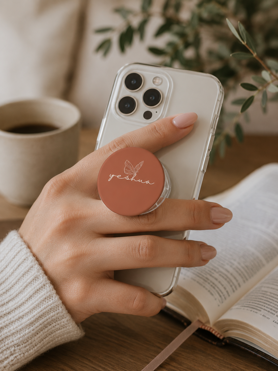 A person holding a smartphone featuring a terracotta-colored pop-grip with a butterfly icon and the word "Yeshua" written in white script.