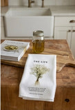 A white waffle-weave kitchen towel featuring a bouquet of dried baby's breath and the text "THE LIFE" laid on a rustic wooden cutting board next to a glass jar and a cookbook.