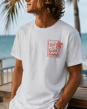 Man wearing a white faith-based t-shirt featuring a red "God is Good" palm tree stamp graphic, styled for a coastal sunset lifestyle.